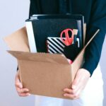 Woman carrying cardboard box with office items like scissors and notebooks.